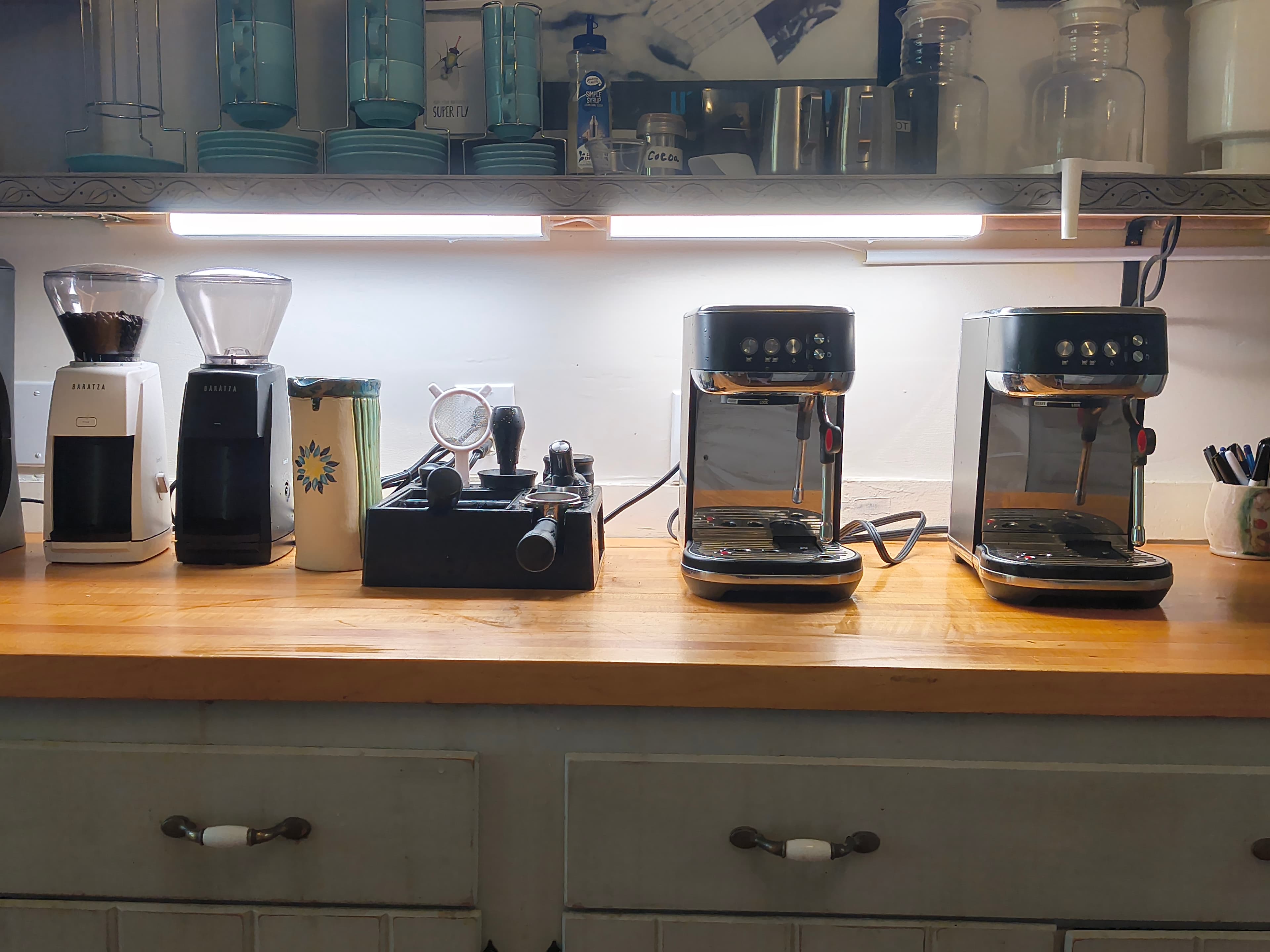 A well-organized kitchen countertop featuring two espresso machines, a coffee grinder, and brewing tools under bright lighting.