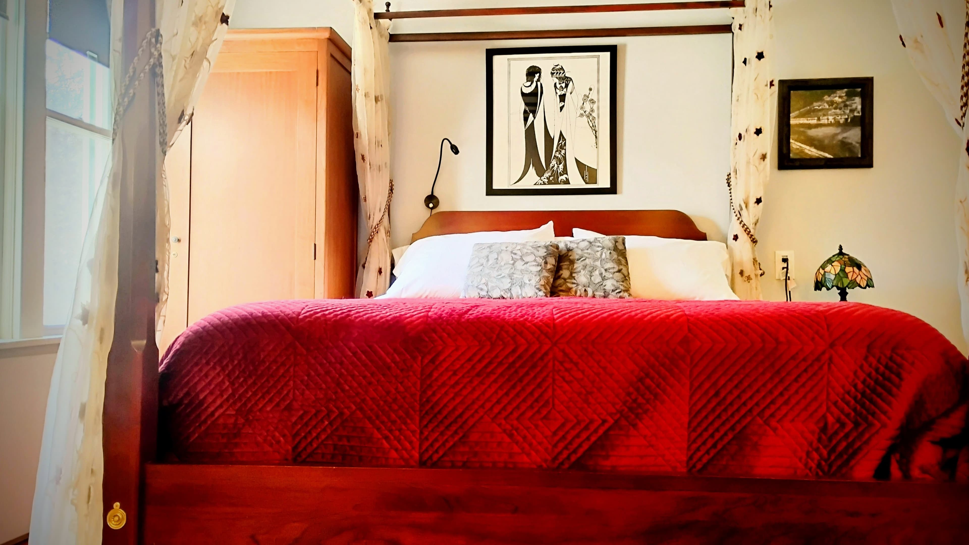 A cozy bedroom featuring a red quilted bed, elegant artwork on the walls, and warm lighting.