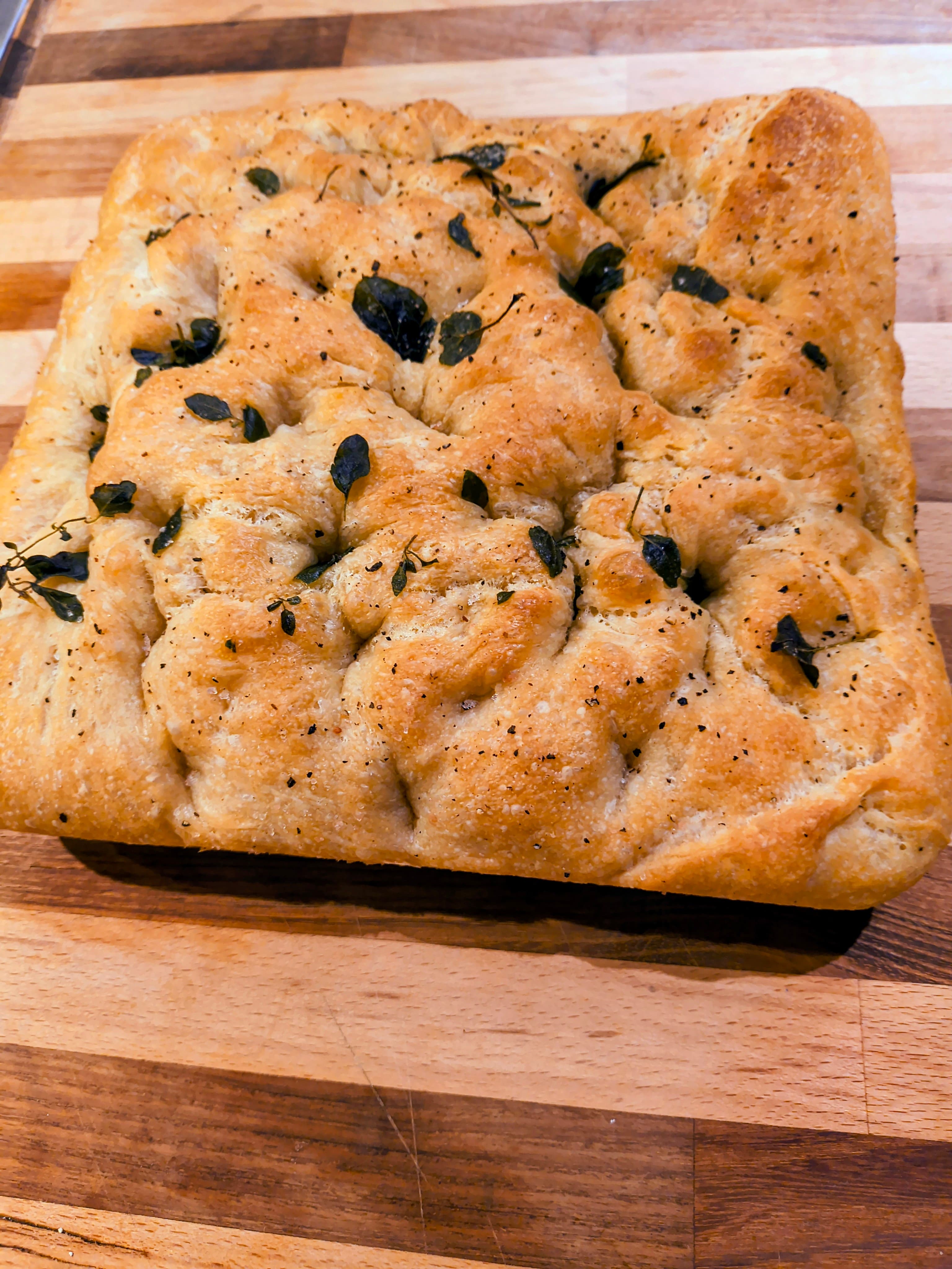 A golden-brown focaccia topped with herbs rests on a wooden cutting board.
