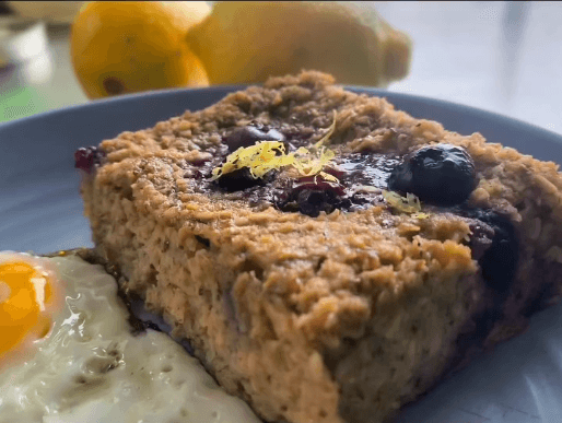 A slice of baked oatmeal topped with blueberries and lemon zest, next to a sunny-side-up egg and two lemons in the background.