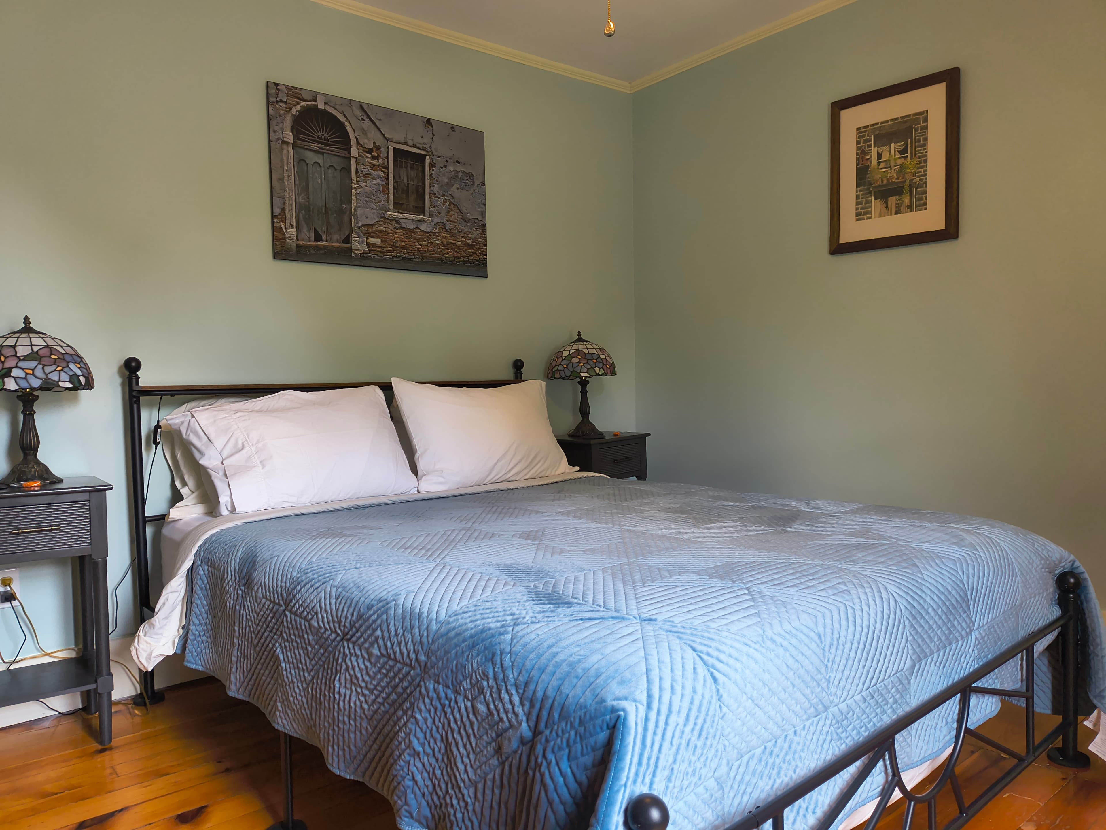 A cozy bedroom featuring a bed with a blue quilt, two bedside lamps, and wall art.